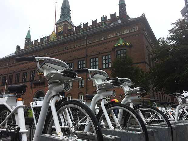 A row of white electric bicycles in front of a stately brick building in Copenhagen.