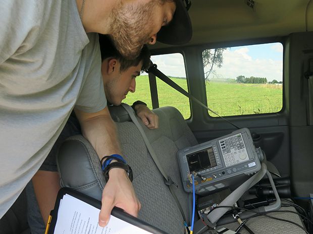 NYU students George MacCartney (left) and Jeton Koka (right) lean over the backseat of a van to observe the signal strength from their receiver on a Keysight E4407B spectrum analyzer, resting on a van seat, before ​recording a measurement. 