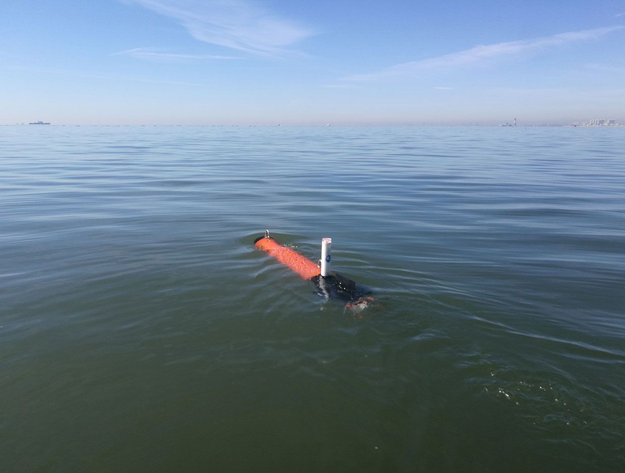 A photo of the top of an orange AUV moving away from the camera in the water on a clear sky day. 