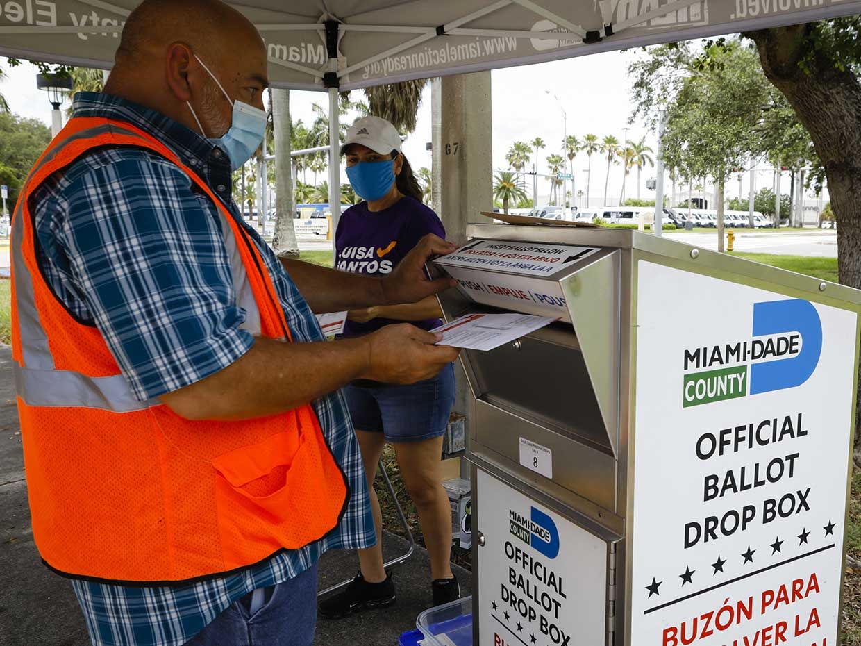 An election official wearing a protective mask drops off a mail-in ballot in a drop box at a polling location in Miami, Florida, U.S., on Tuesday, Aug. 18, 2020. 