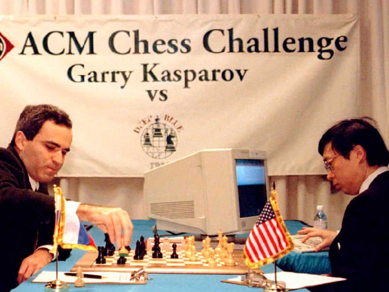 Photo of a man playing chess and a man sitting next to a computer.