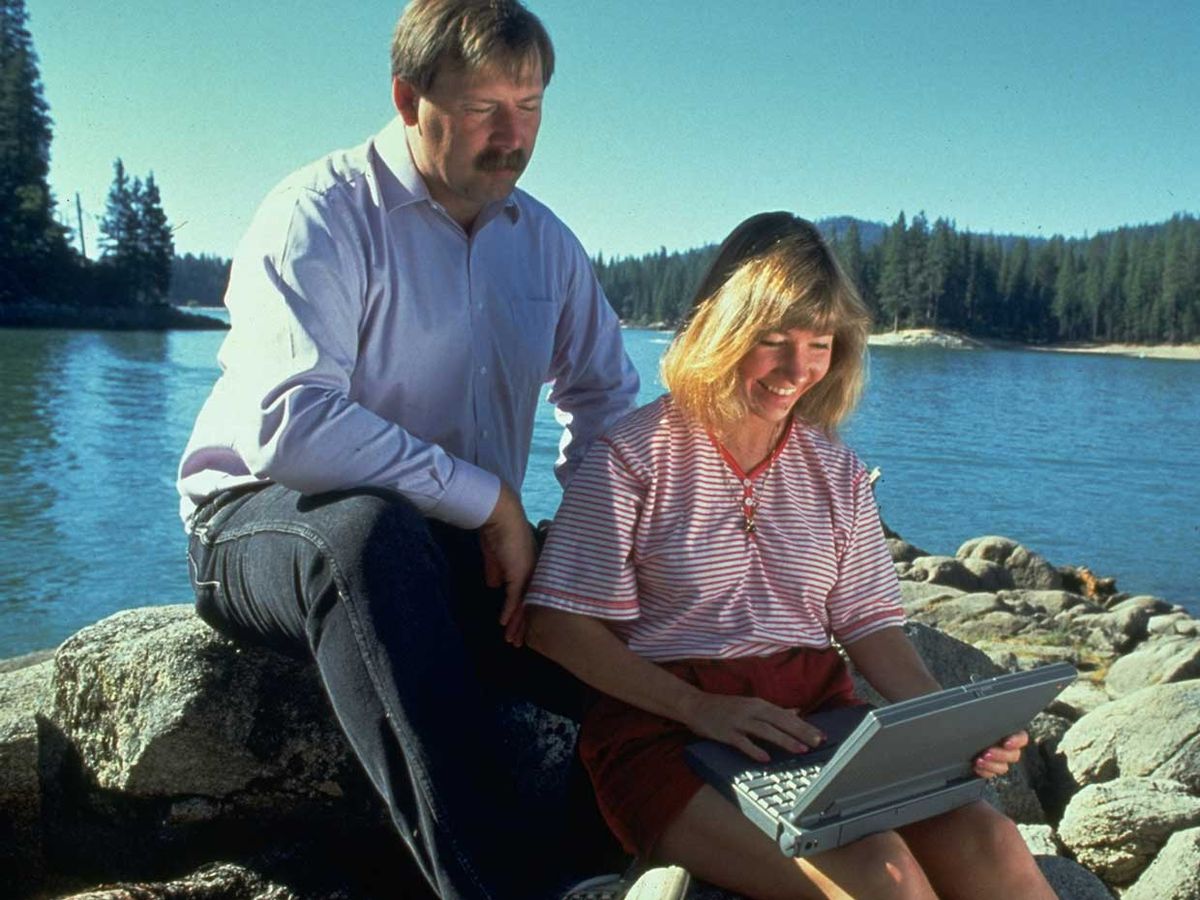 1993 photo of Sierra Systems founders Ken & Roberta Williams sitting on rocks on lakeshore using laptop computer.