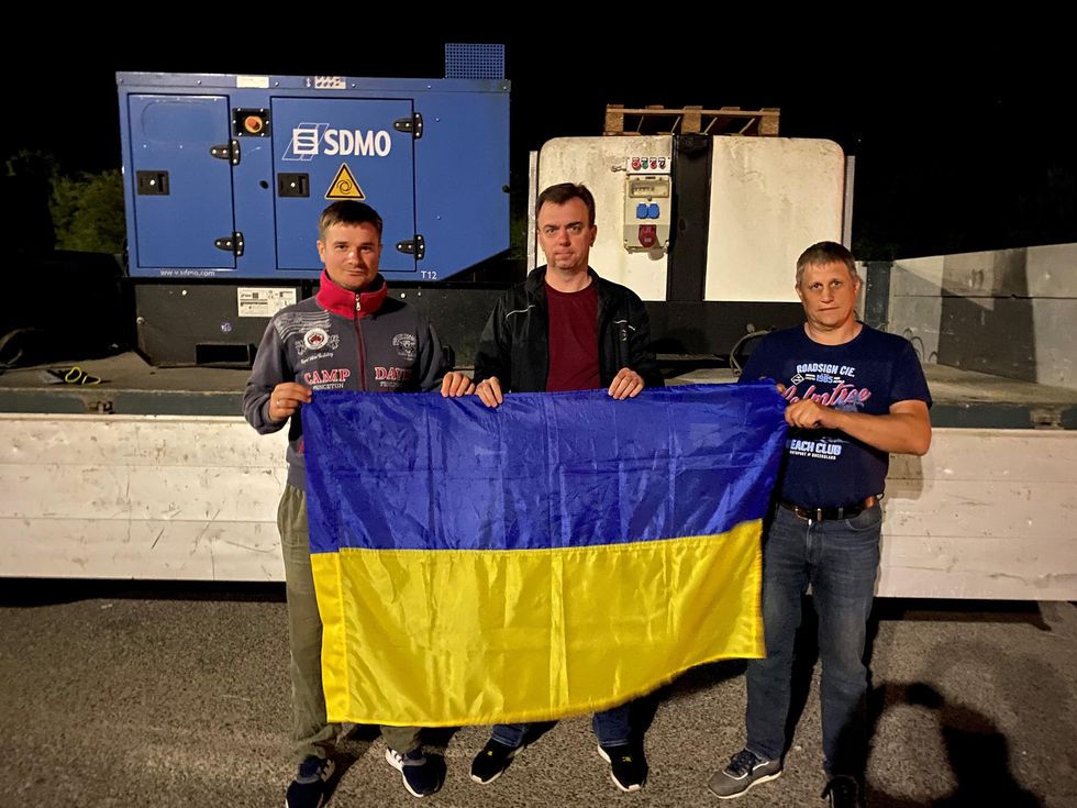 3 men hold a Ukrainian flag in front of electrical equipment