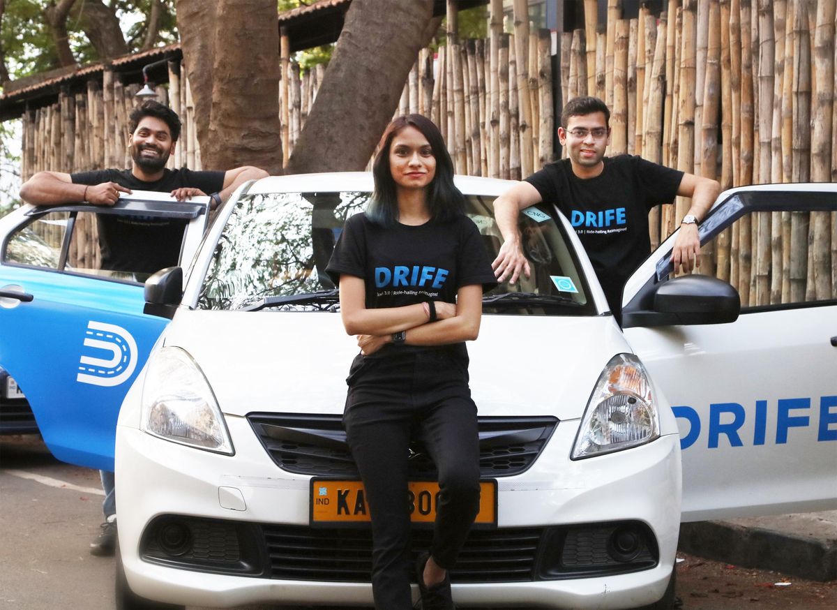 3 people stand at a car with the Drife logo on it smiling at the camera