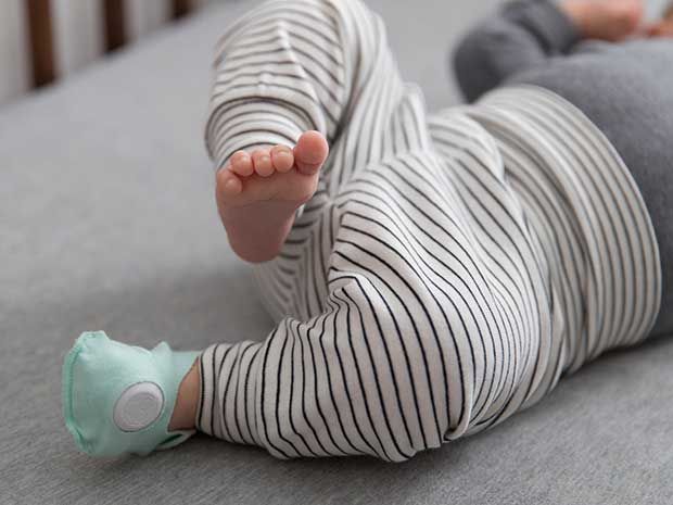 A baby lying in a crib is wearing one green cotton sock with embedded electronics.