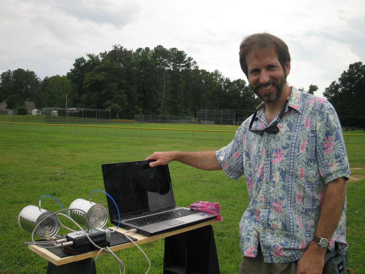 A bearded smiling man in a Hawaiian shirt stands in a field, in front of a workbench, on which sits a laptop, and a contraption with two aluminum cans with wires coming out of them connected to an electronic device.