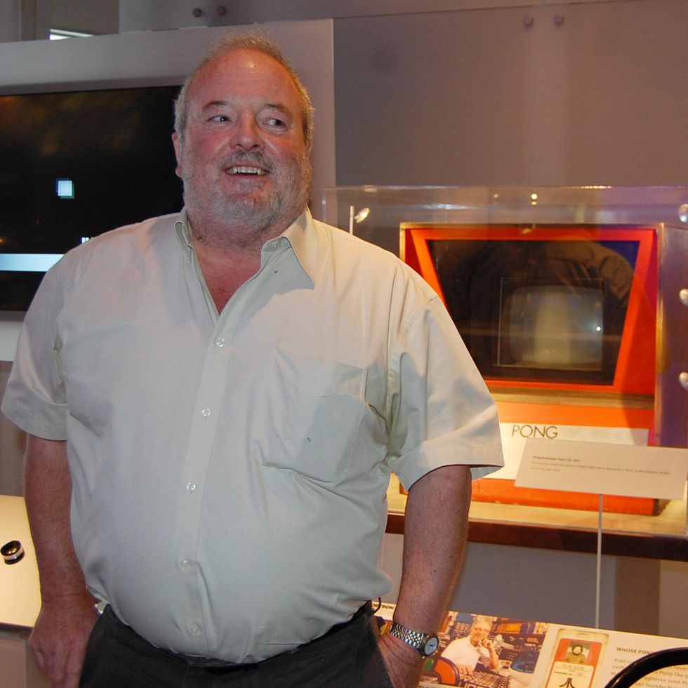 A bearded white man smiles as he stands in front of a retro game console.