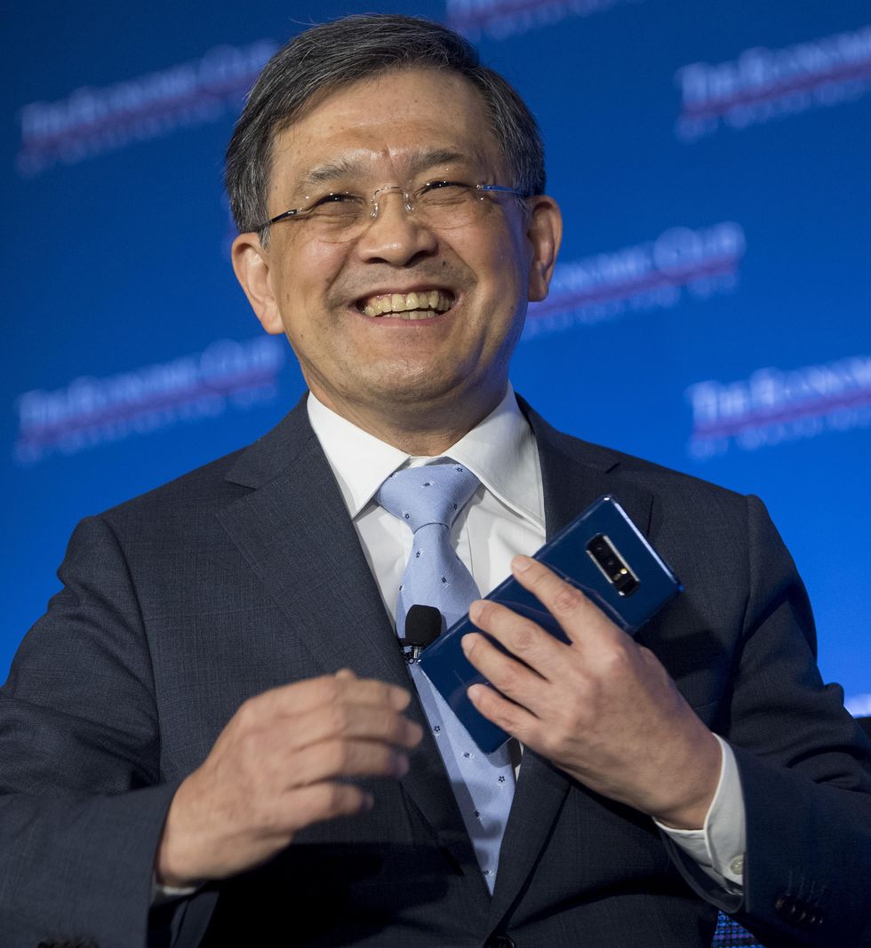A bespectacled man in a suit smiles while holding a smartphone.
