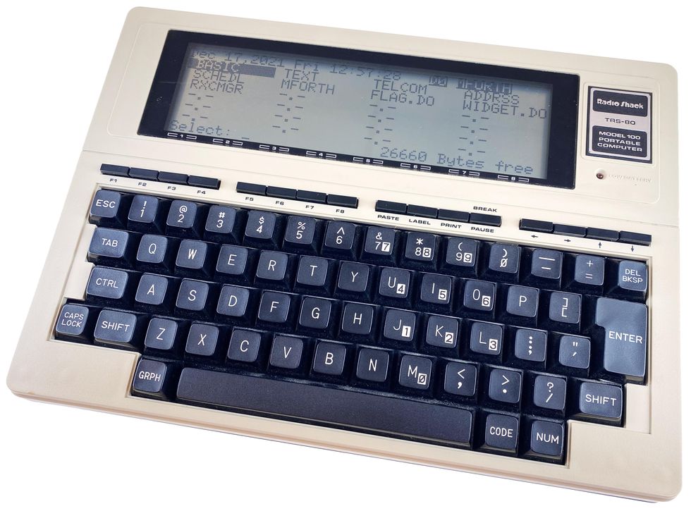 A black and white book-sized computer with a keyboard and monochrome LCD screen labeled "Model 100 Portable Computer"