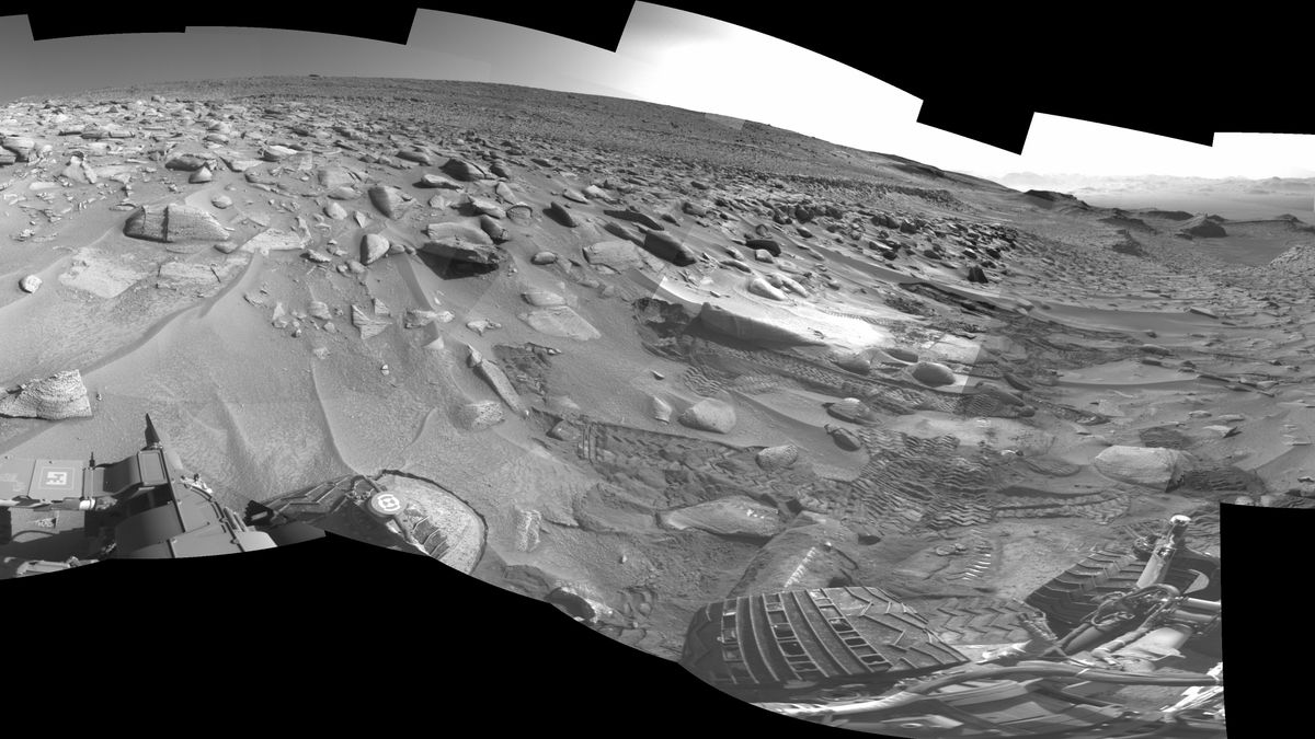 A black and white composite photo showing rocky, dusty, steep terrain on Mars with the wheels of a robotic rover visible in the bottom corner