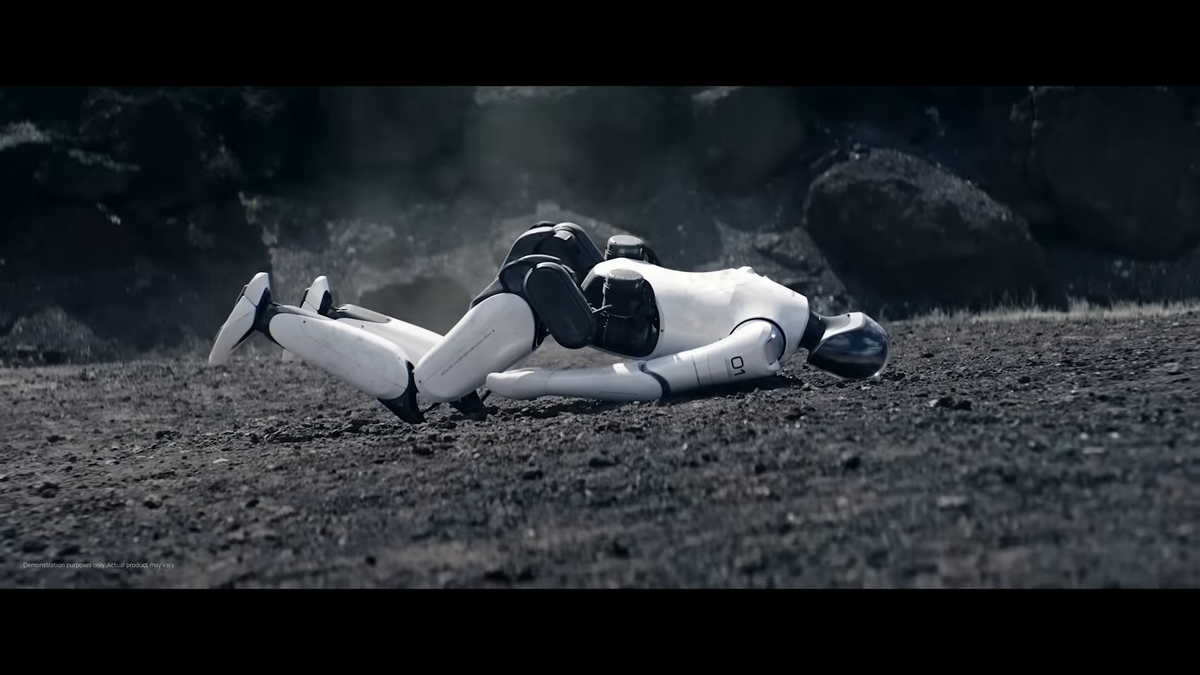 A black and white humanoid robot lies face down on dirt after appearing to have just fallen