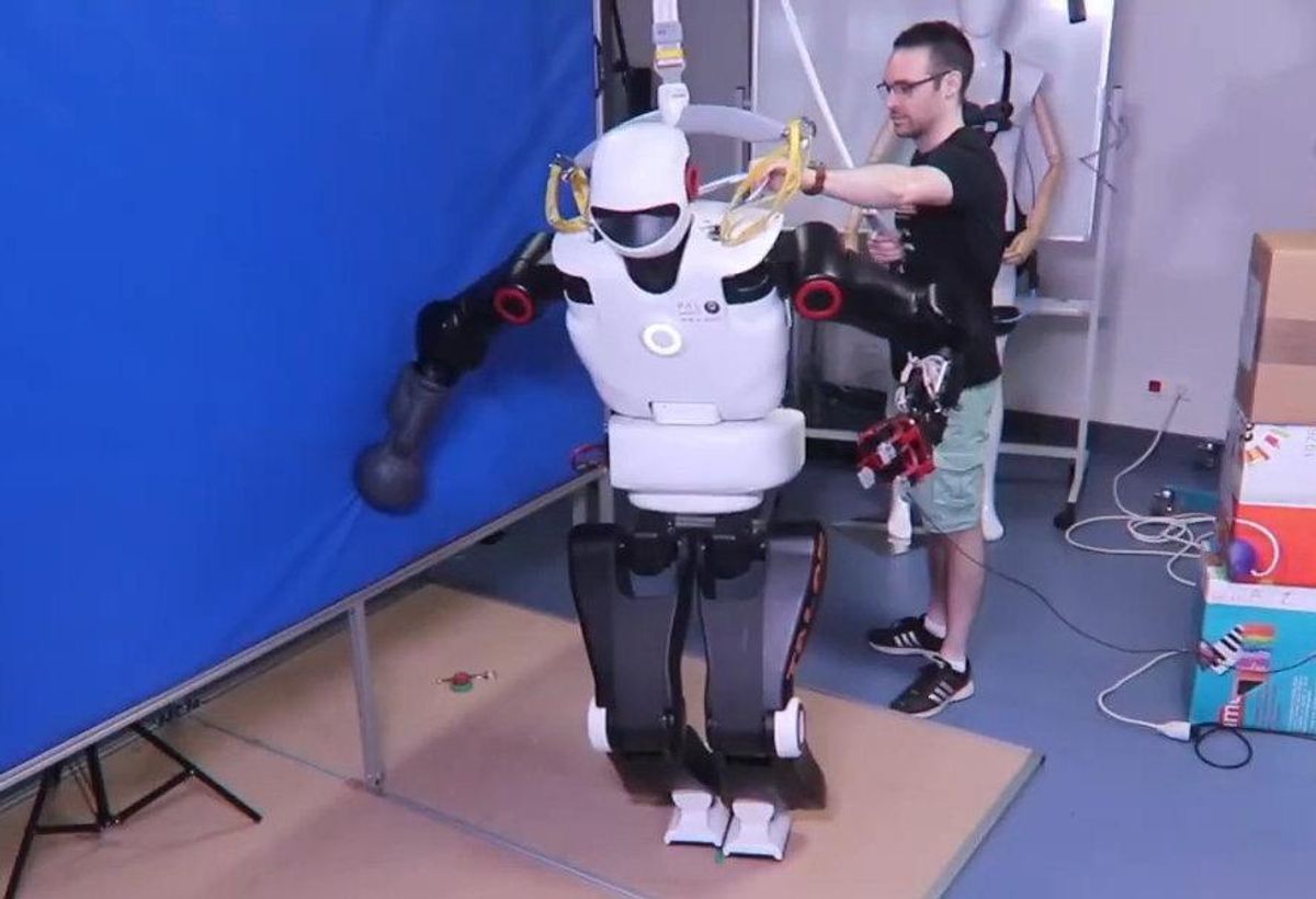 A black and white humanoid robot with a malfunctioning leg supports itself with one arm against a wall