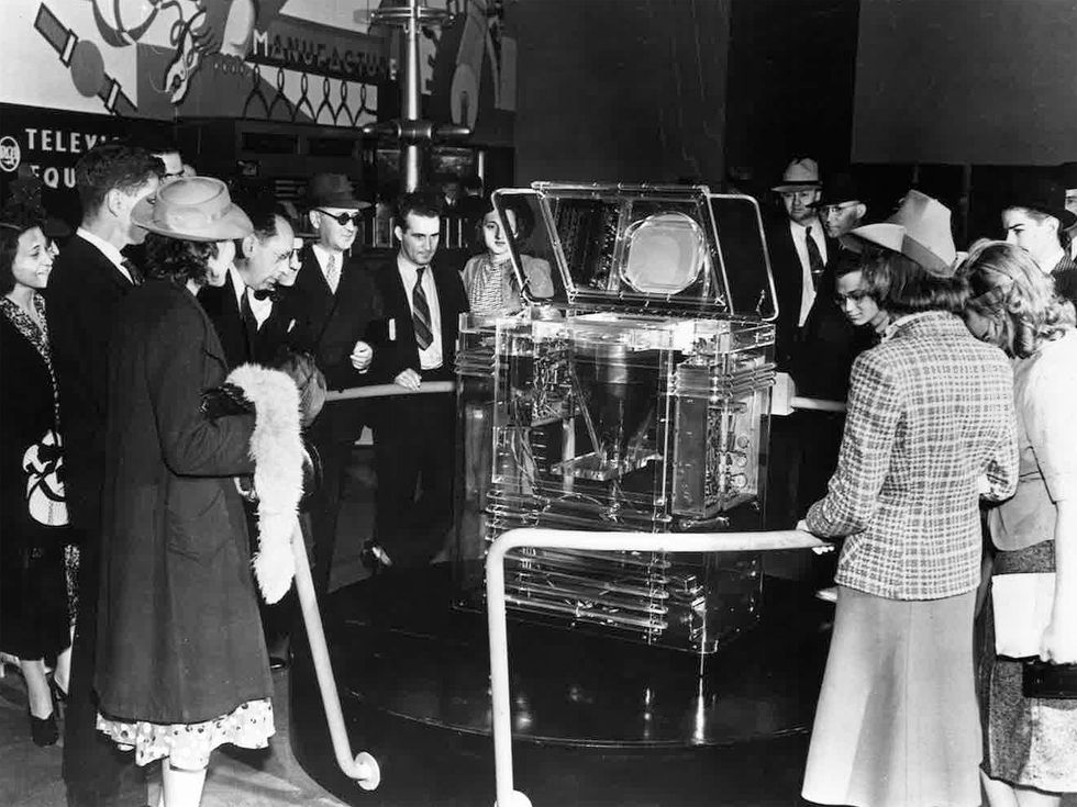 A black and white photo of a crowd of people gathered around a lucite television set.