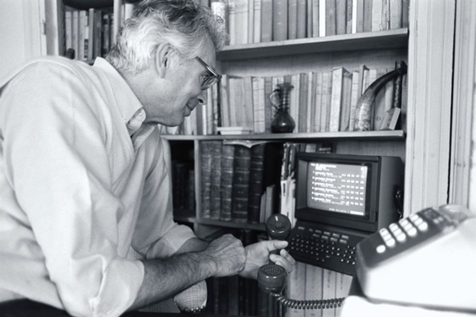 A black and white photo of a man holding a phone and looking at a small monitor on a shelf. 