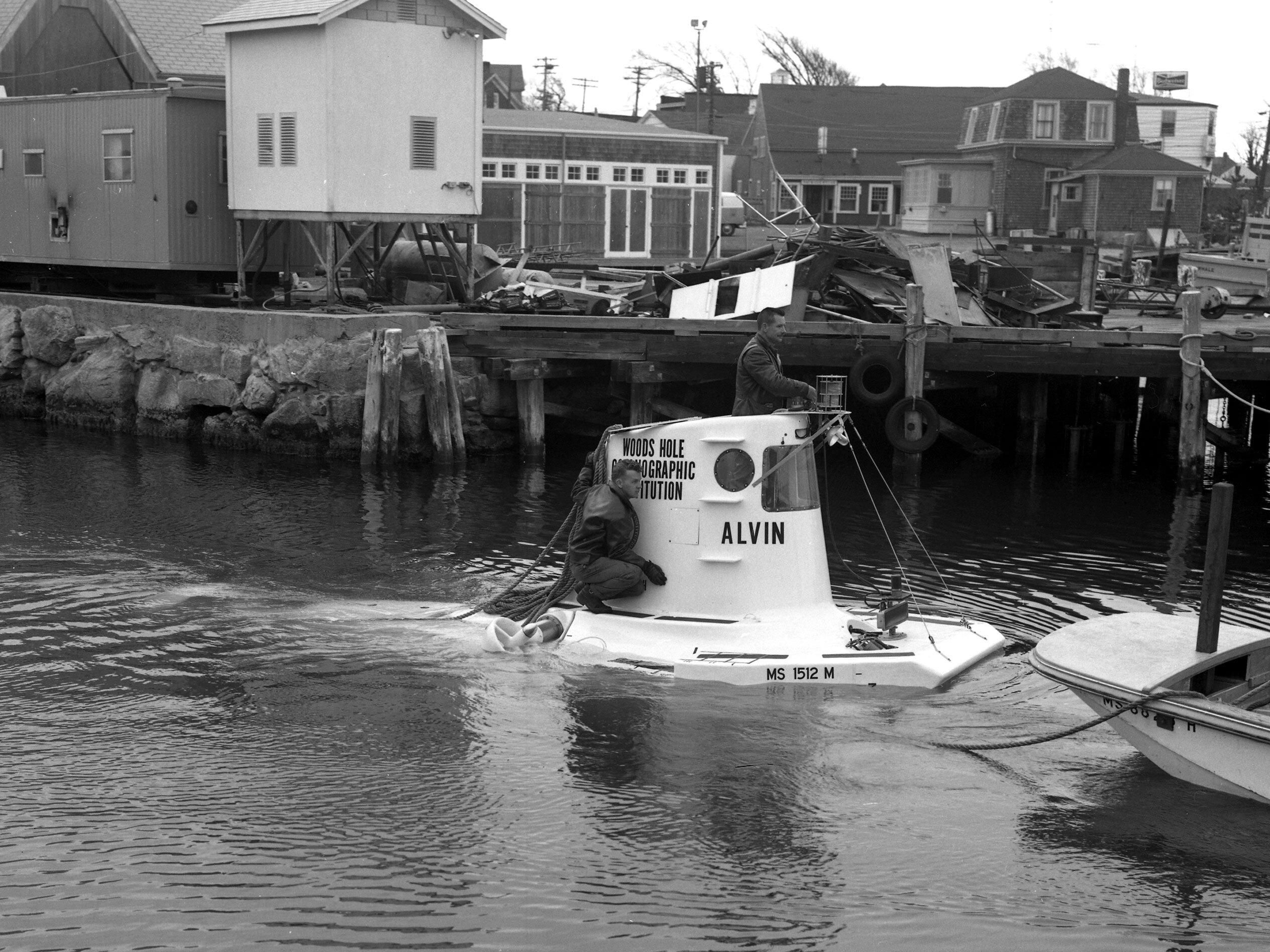 A black and white photo of a submersible in the water in front of a pier with debris on it.  