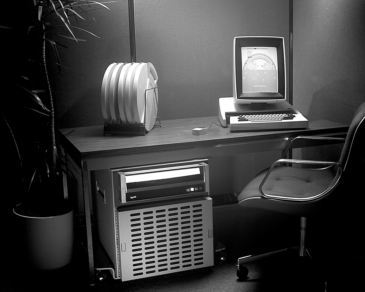 A black and white photo of a vertical computer display and keyboard and mouse on a desk, with the computer itself under the desk.  