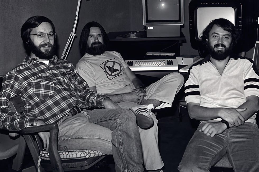 A black and white photo of three bearded men in casual clothes sitting in front of old computers.