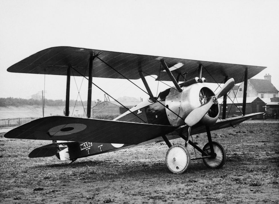 A black-and-white photo shows a single-seat, two-wing plane with a silver front end carrying a propeller. The plane, which is parked on an open field, has two large white wheels in the front.
