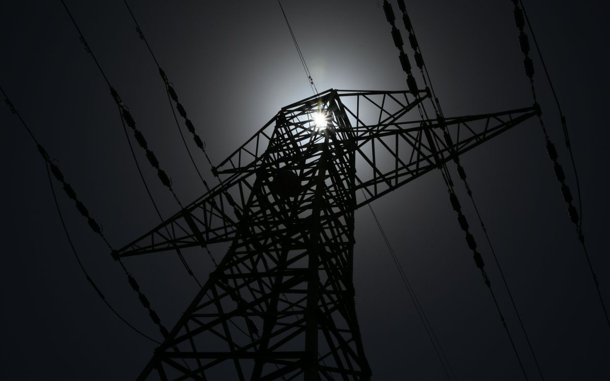 A black and white photograph shot from below of an electricity pylon that is dark except for a ray of sun coming through the top.