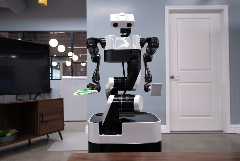 A black and white two armed robot with a mobile base sweeps the floor of a living room