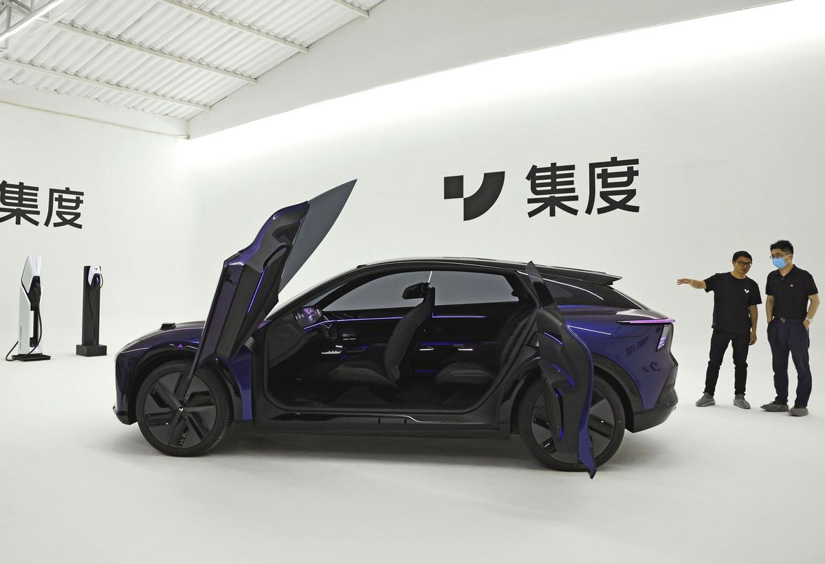 A black car sits against a white backdrop decorated with Chinese writing. The car’s doors are open, like a butterfly’s wings. Two charging stations are on the car’s left; two men stand on the right.