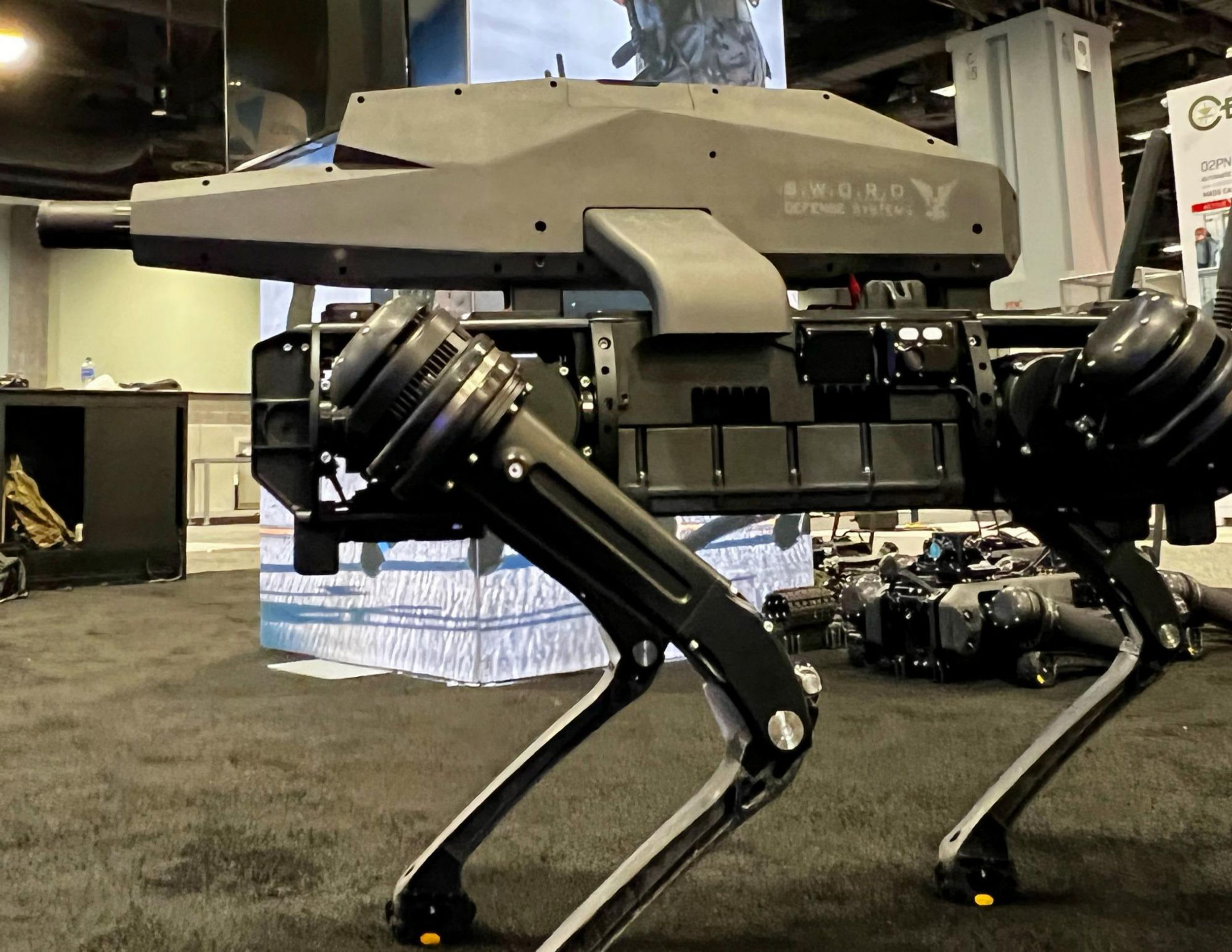 A black quadrupedal robot with a futuristic rifle on its back stands in an exhibition hall