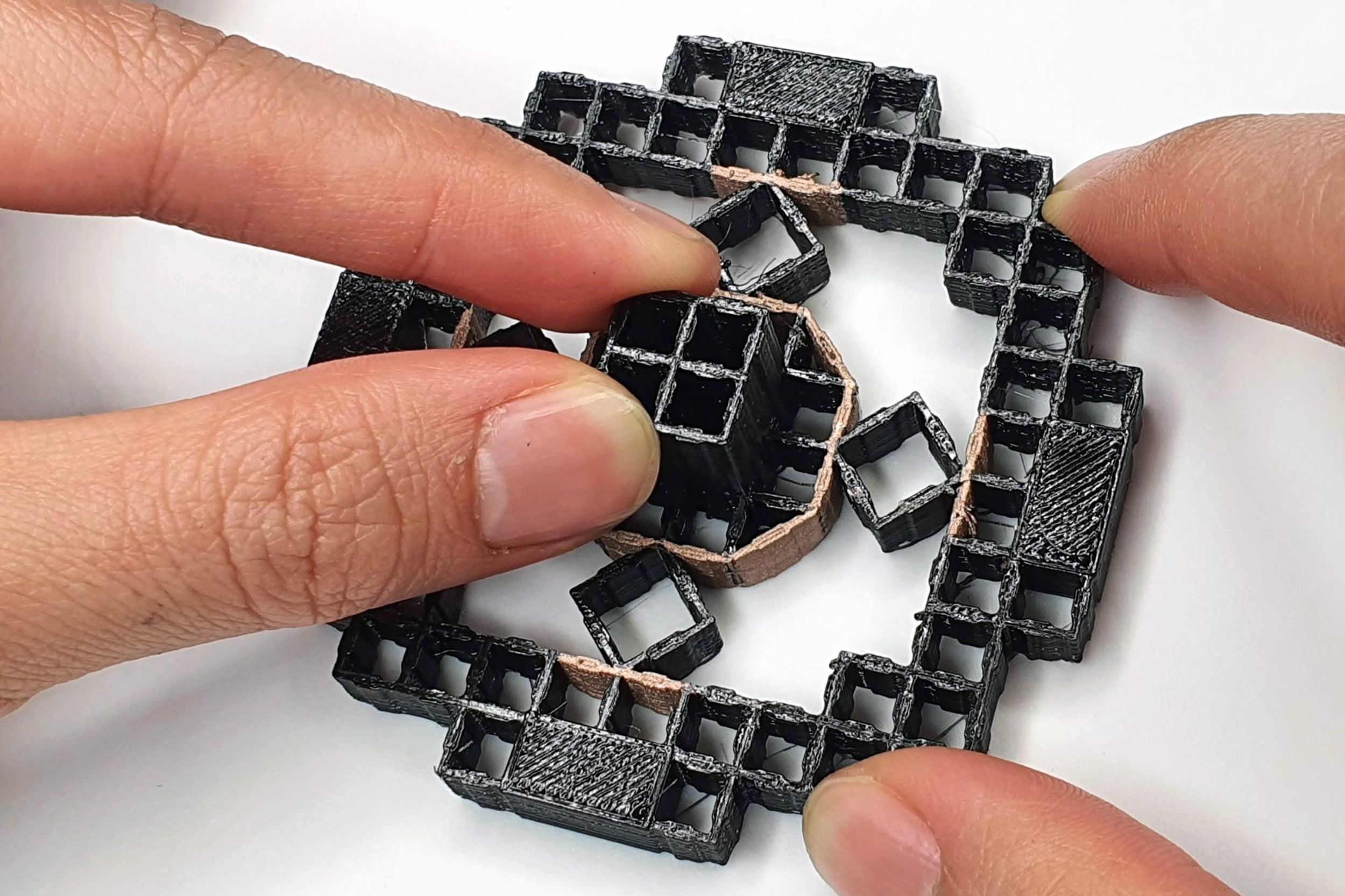 A black shiny structure composed of a grid of squares forming a larger square. The center is a gold circular piece with more squares, and 4 squares on the top, bottom, left, and right. 4 fingers are holding it to show the scale being about a finger length.