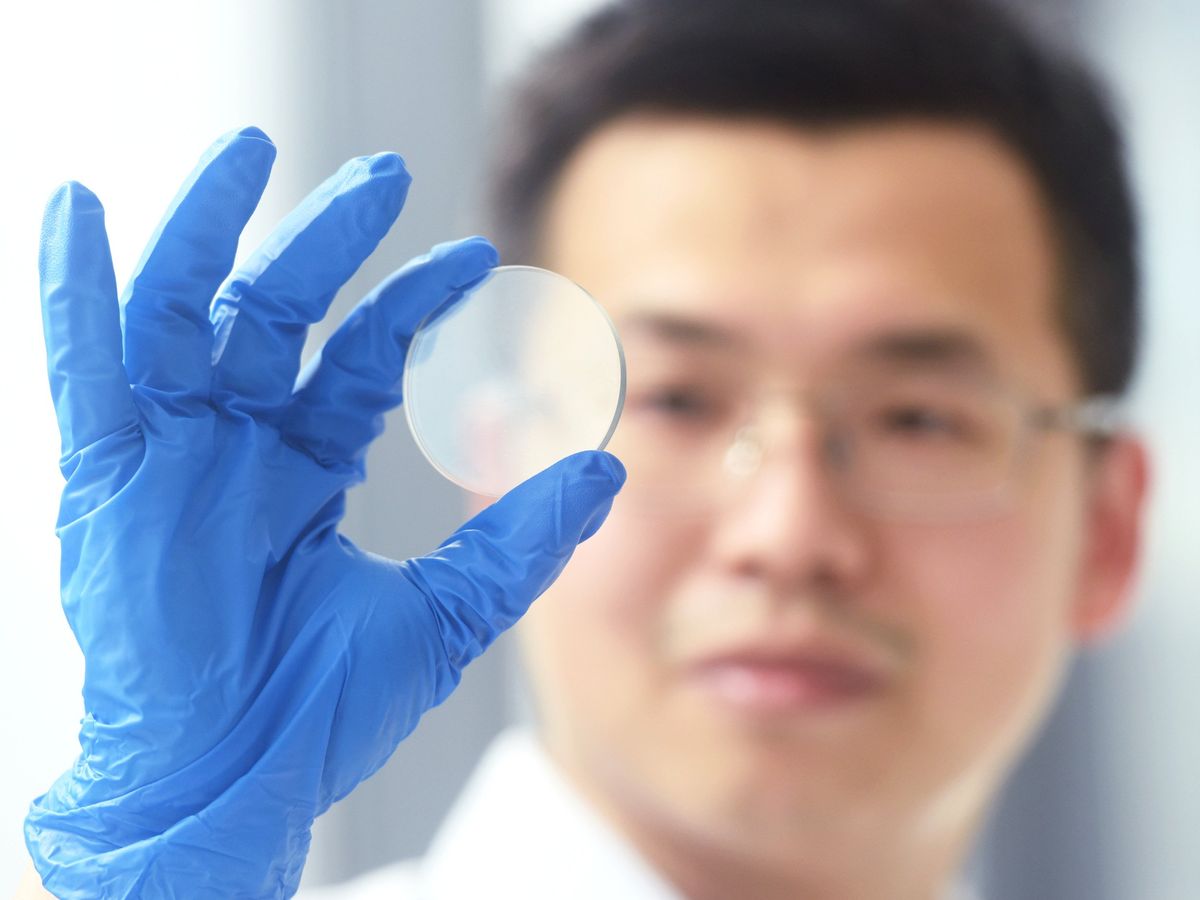 A blue gloved hand holds up a translucent disc. An out of focus face is in background.