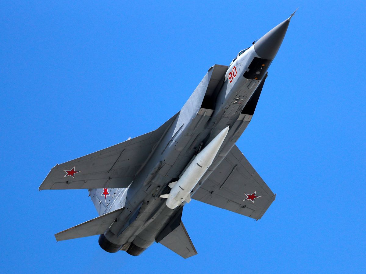 A blue-gray, twin-engine fighter jet is seen from below, against a clear, blue sky; a red star is visible on the underside of each wing and on the side of the vertical stabilizer. Carried under the fuselage is a white rocket with small fins at the back.