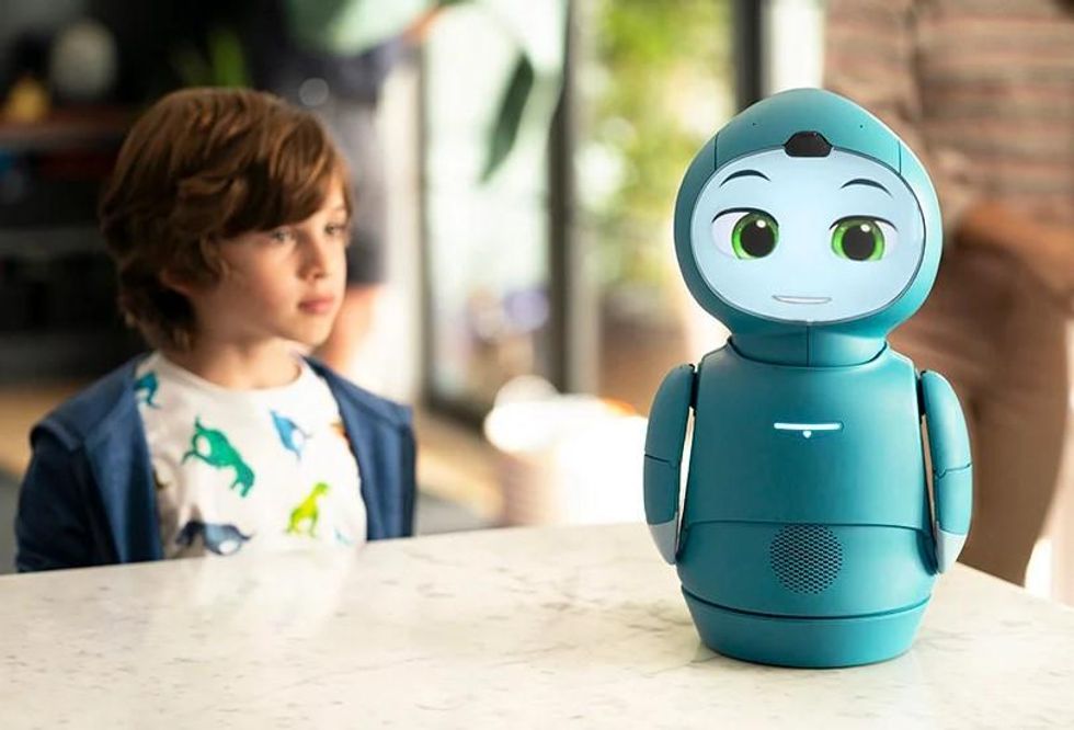 A boy with brown hair observes a Moxie robot, which has green body the size of a coffee maker with a screen as a face showing green eyes, eyebrows, and a mouth.