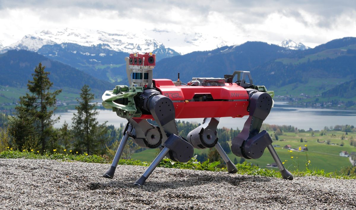 A bright orange four legged robotic dog stands on a grave patch at the top of a mountain with Swiss countryside in the background