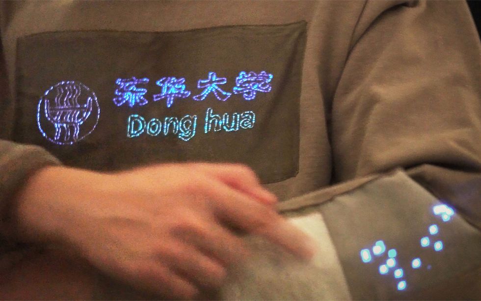A brown shirt with Dong hua and Chinese lettering on it in glowing blue thread.