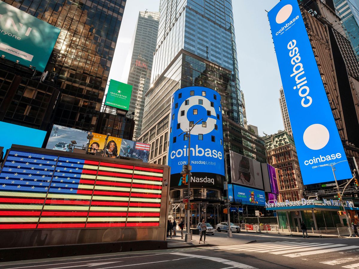 A busy city intersection with a large screen showing an American flag, and two other showing the word Coinbase, and its logo.