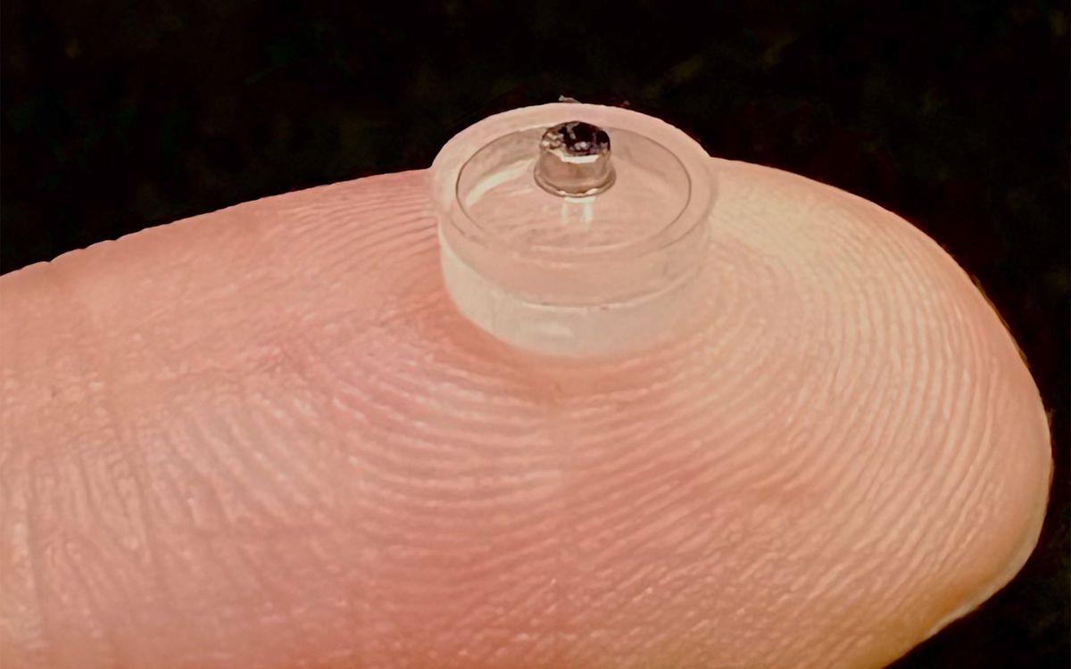A clear circular object with a metal protrusion sits on the tip of a persons finger.