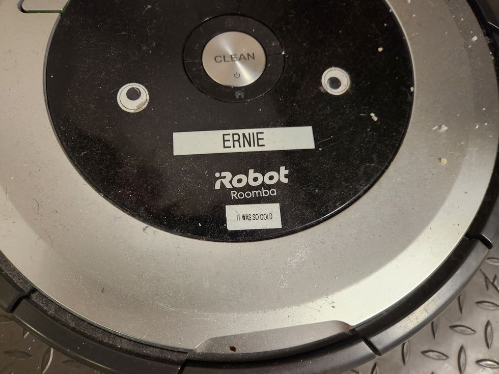 A close up picture of  the top of a Roomba showing some small eye stickers, a sticker with the name "Ernie," and a sticker that says "it was so cold."