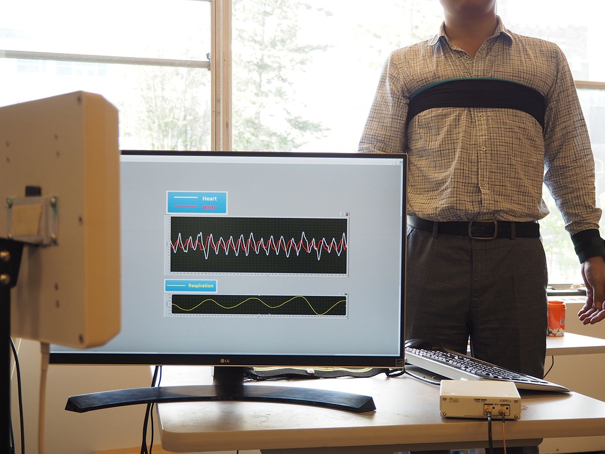 A computer and equipment in the foreground show the monitoring of vital signs for a patient in the background with black straps around his chest and wrist.