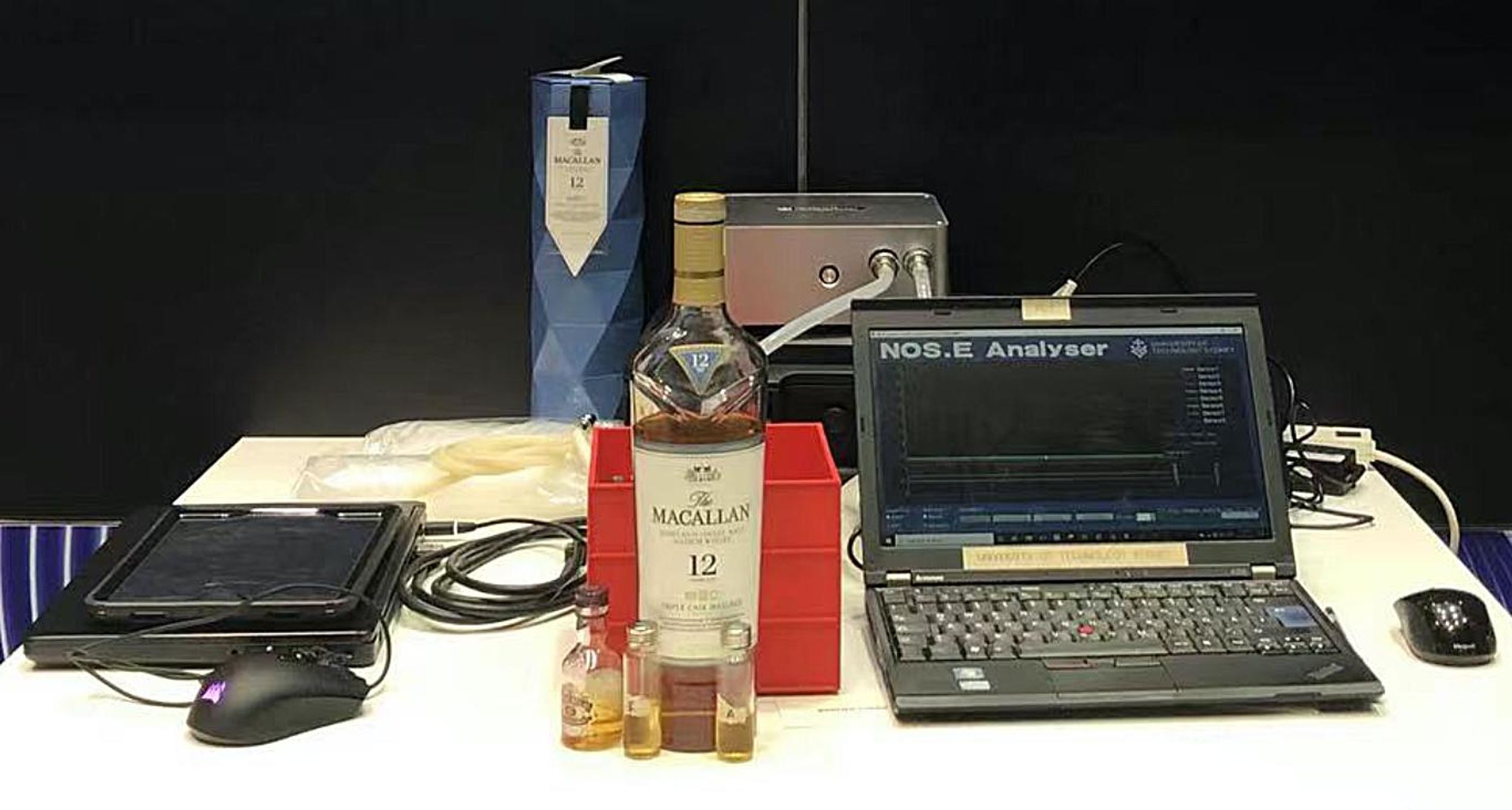 A computer is hooked up to equipment. Multiple bottles of whiskey large and small sit on the table.