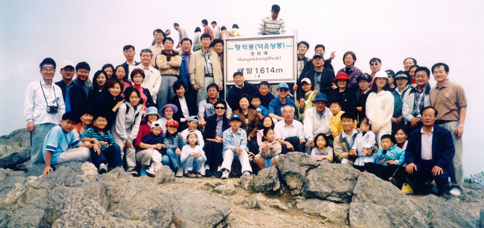 A crowd of more than two dozen people seated and standing atop a rock formation. A large white sign is at center.