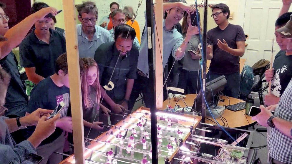 A crowd of people including two children surround a brightly lit foosball table. A man and girl play on one side, while electromechanical machinery plays on the other.