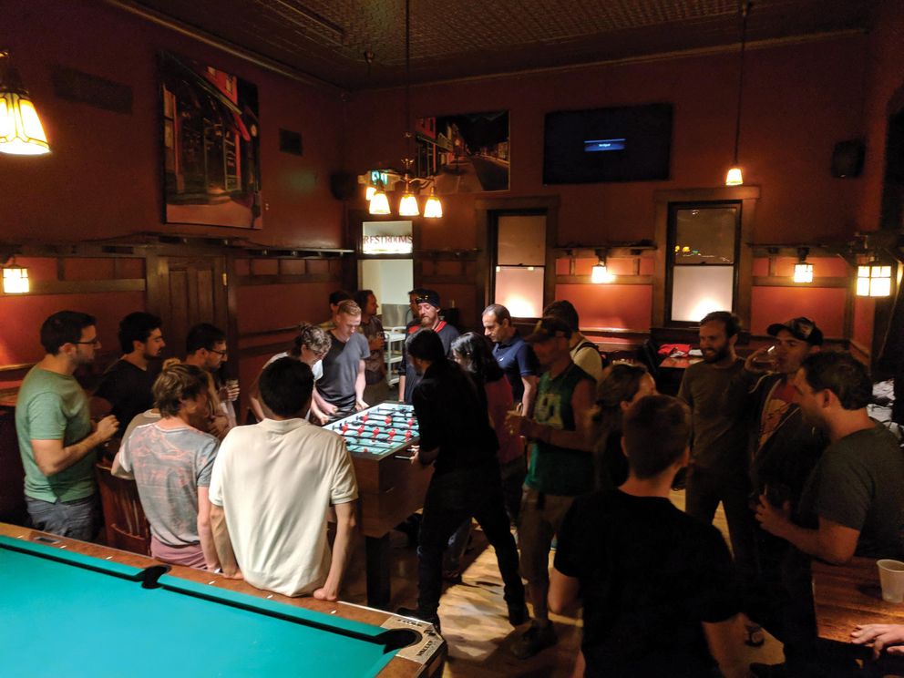 A crowd of people surround a foosball table in a dimly-lit bar. 