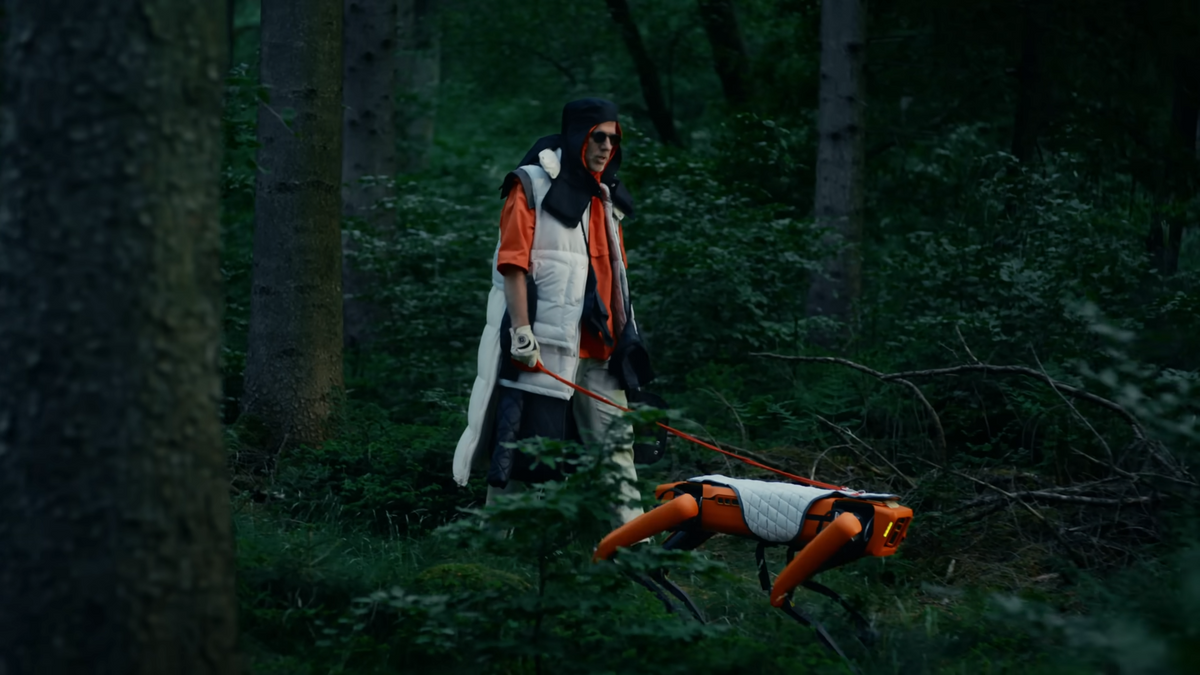 A curiously dressed man wearing white and orange clothes and dark glasses walks an orange robot dog in a white coat through the woods