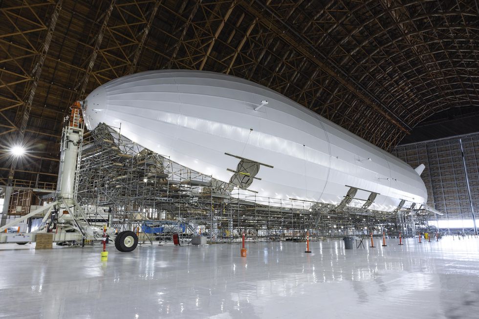 A cylindrical white airship under construction in a large aircraft hanger.