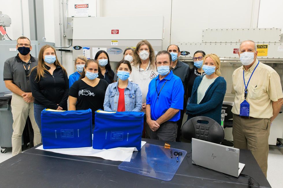 A dozen researchers in masks stand in front of two blue bags in a NASA laboratory
