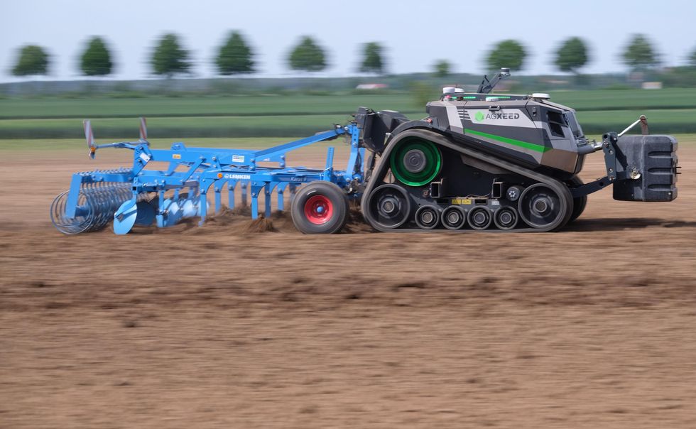 A driverless farm tractor uses one of the implements mounted on its front to till soil autonomously.
