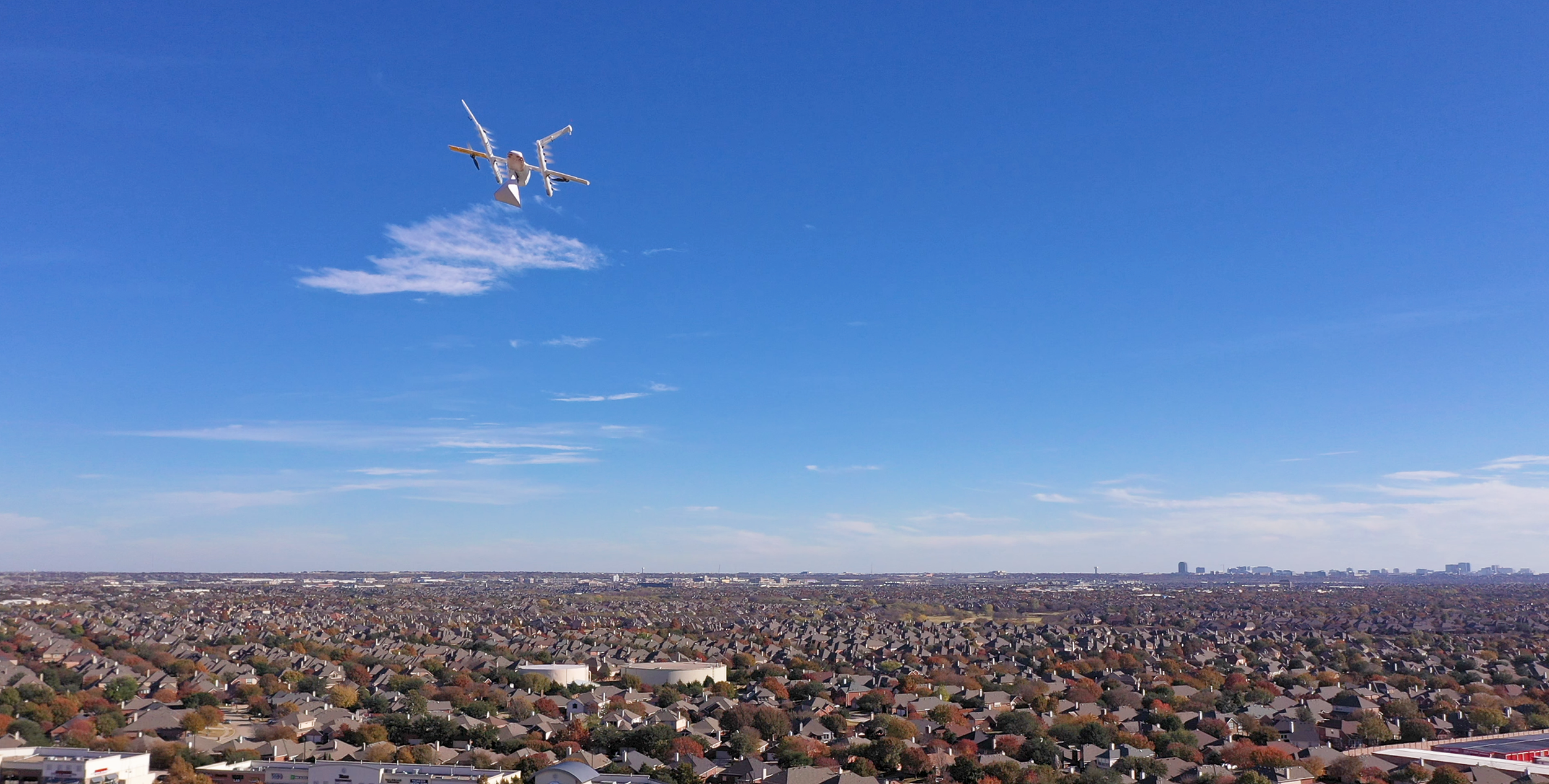 A drone carrying a package flies high above a flat suburban landscape