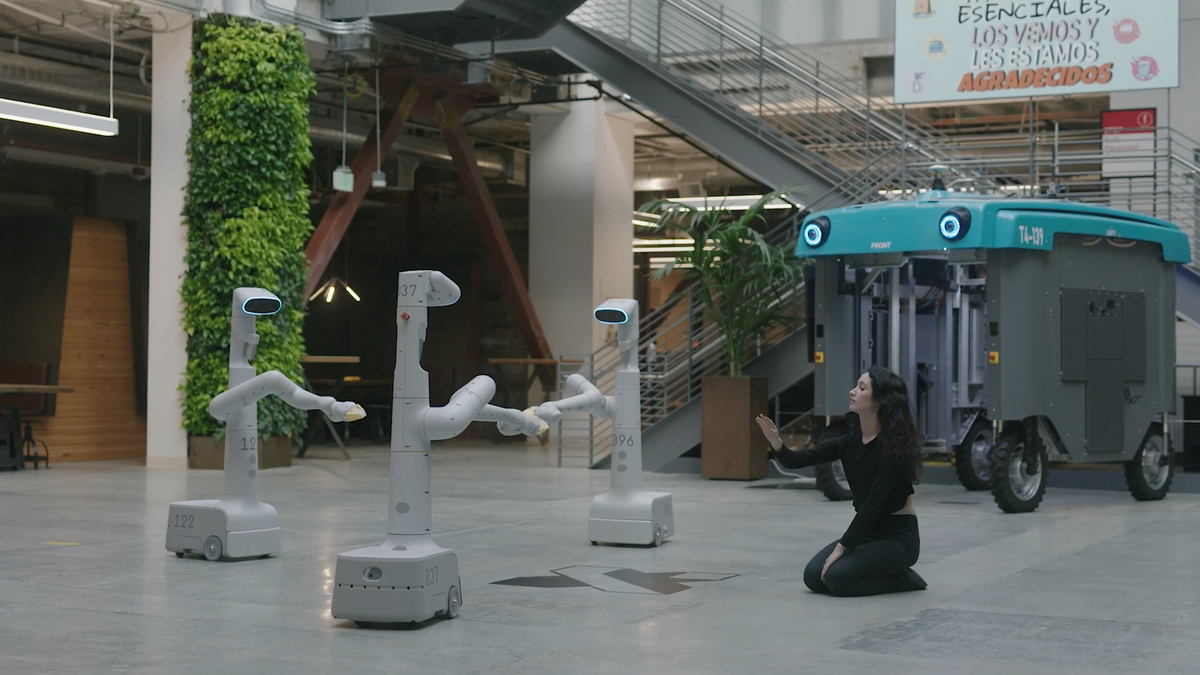 A female dancer dressed in black interacts with three white mobile robots with heads and single arms