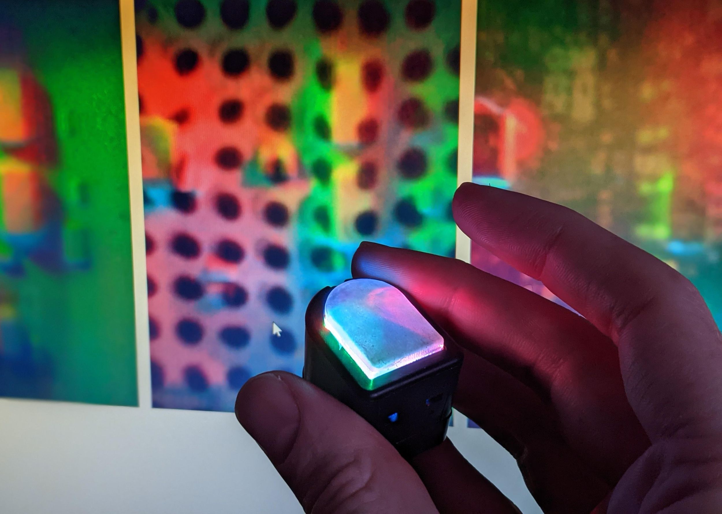 A fingertip-sized tactile sensor glowing with red, blue, and green LEDs held between finger and thumb