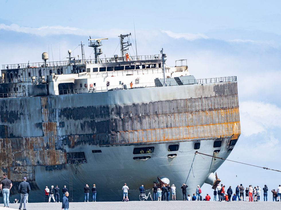 A fire-ravaged ship is docked at a port while workers and onlookers examine the damage