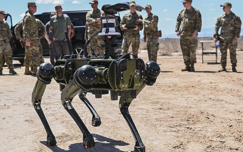 A four-legged black and grey robot moves in the foreground, while in the background a number of uniformed people watch its actions, 