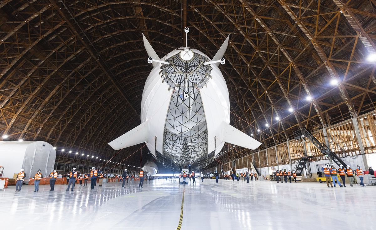 A giant white airship in a hangar, with many people below it to show its large scale.